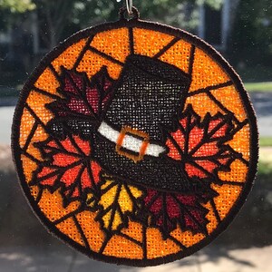 May include: A round, orange, brown, and red embroidered ornament with a pilgrim hat and fall leaves. The ornament is hanging from a suction cup.