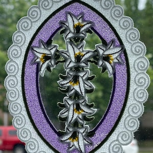 May include: A white cross with white flowers is embroidered on a purple oval background. The oval is surrounded by a white lace border.