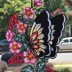 May include: A black and white butterfly with yellow and red accents is embroidered on a white fabric with green leaves and pink and red flowers.