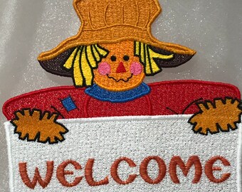 May include: A scarecrow with a red shirt, blue collar, and yellow straw hat holds a white sign that says "WELCOME".