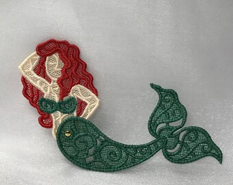 May include: A green and red embroidered mermaid with a gold bead accent. The mermaid is in a sitting position with her tail curled behind her.