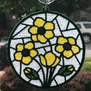 May include: A round, white, embroidered window hanging with a green border and three yellow flowers with black centers.