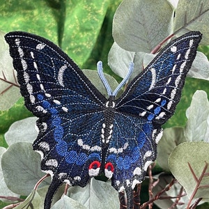 May include: A blue and black embroidered butterfly patch with white accents and red eyes. The butterfly is on a green and gray background.