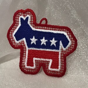 May include: Red, white, and blue embroidered donkey ornament with a white mesh background. The donkey has three white stars on its chest.