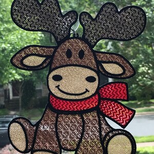 May include: A brown moose with a red scarf, sitting with a big smile. The moose is made of a sparkly material and has a black outline.