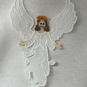 May include: White embroidered angel with a halo and brown hair. The angel is standing with its wings spread out.