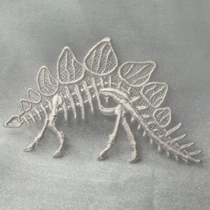May include: White embroidered patch in the shape of a Stegosaurus dinosaur. The patch features intricate lace-like detailing, including the dinosaur's plates, legs, and tail. The patch is likely for clothing or craft projects.