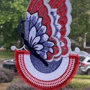 May include: A red, white, and blue embroidered butterfly with a heart charm hanging from the bottom. The butterfly is hanging from a window with a suction cup.