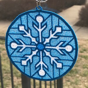 May include: A blue and white snowflake ornament with a suction cup for hanging on a window. The snowflake is embroidered with a white outline and a blue center.