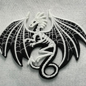 May include: White embroidered skeleton dragon with black wings and a web pattern. The dragon is curled up with its wings spread out.