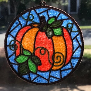 May include: A round, embroidered pumpkin ornament with a stained glass design. The pumpkin is orange with green leaves and vines. The background is blue.