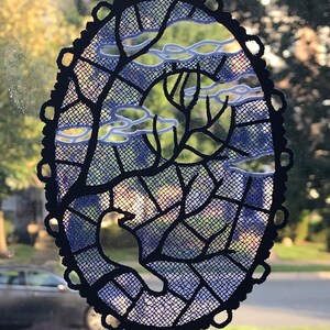 May include: A black and blue oval-shaped lace ornament with a tree design and white clouds. The ornament is hanging from a window.