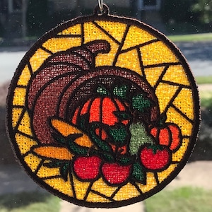 May include: A round, embroidered, stained glass-style ornament featuring a cornucopia overflowing with pumpkins, gourds, and tomatoes. The ornament is decorated with a yellow, brown, orange, green, and red color scheme.
