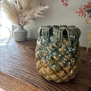May include: A ceramic pineapple-shaped vase with a green and brown glaze. The vase has a textured surface and is sitting on a wooden shelf.