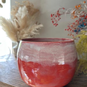 Puede incluir: Un macetero de cerámica rojo y blanco con un acabado brillante. El macetero tiene una forma redondeada y un esmalte blanco que se funde en un esmalte rojo.