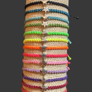 May include: A display of colourful friendship bracelets with star charms. The bracelets are arranged on a burlap-covered cylinder. The bracelets are in various colours, including black, blue, green, yellow, orange, pink, purple, and red.