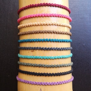 May include: A collection of 12 colourful braided bracelets displayed on a wooden surface. The bracelets are made of a variety of colours, including red, pink, beige, brown, teal, grey, turquoise, green, black, and purple.