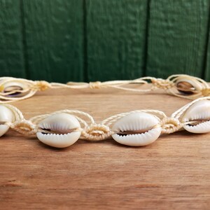 May include: A white shell necklace with a knotted cord. The necklace is made with cowrie shells and has a simple design.