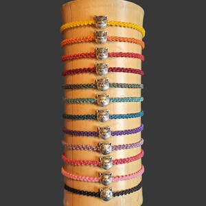 May include: A display of 12 colourful braided bracelets with silver cat head charms. The bracelets are arranged in a rainbow pattern on a wooden stand.
