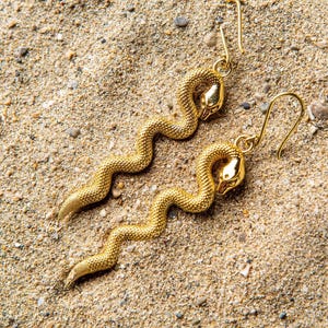 Könnte beinhalten: Ein Paar goldene Schlangenohrringe mit strukturierter Oberfläche. Die Ohrringe hängen und haben einen Hakenverschluss.