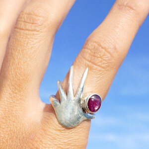 May include: A silver ring with a stylized bird design and a pink gemstone. The ring is worn on a finger against a blue sky background.