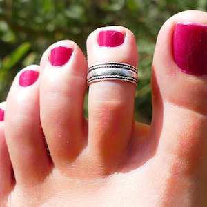 Puede incluir: Un anillo de dedo del pie de plata con una banda texturizada. El anillo se lleva en el segundo dedo del pie de un pie con esmalte de uñas rosa.