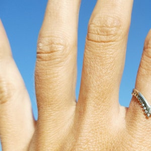Op de afbeelding: Een zilveren ring met een V-vormig ontwerp en kleine decoratieve kralen. De ring wordt gedragen aan een vinger tegen een blauwe lucht achtergrond.