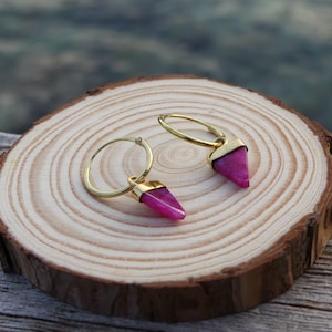 May include: Gold-coloured hoop earrings, each with a magenta triangular gemstone pendant. The earrings are displayed on a wooden slice. The gemstones are pointed and set in gold-coloured metal. The earrings are a stylish accessory.