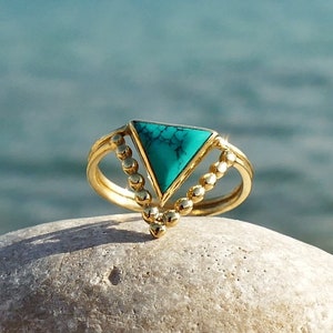 Puede incluir: Un anillo de oro con una piedra preciosa triangular turquesa engastada en el centro. El anillo tiene un diseño de cuentas alrededor de la piedra.