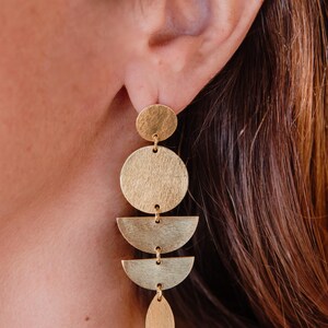 May include: Gold-toned geometric dangle earrings. The earrings feature a series of connected shapes: a small circle, a large circle, two half-circles, and a teardrop shape. The earrings are designed to hang from the ear.