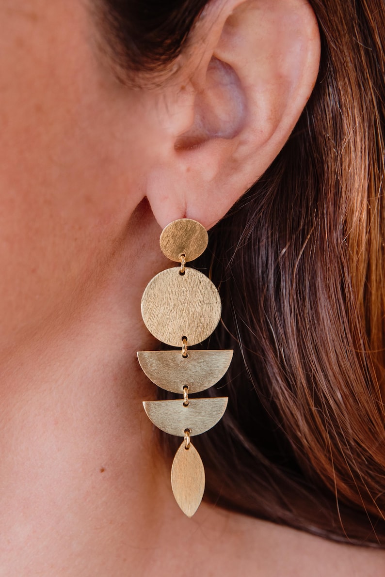 May include: Gold-toned geometric dangle earrings. The earrings feature a series of connected shapes: a small circle, a large circle, two half-circles, and a teardrop shape. The earrings are designed to hang from the ear.