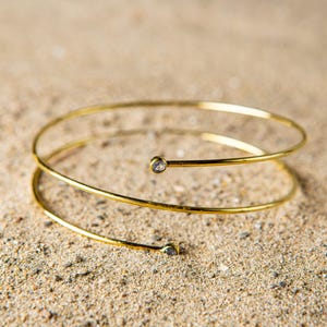 Puede incluir: Un brazalete dorado con un diseño en espiral. El brazalete presenta dos pequeñas gemas transparentes. La joya se exhibe sobre una superficie arenosa, creando un contraste entre el metal y el fondo natural.