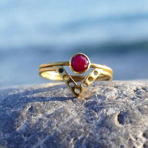 May include: A gold ring with a red gemstone set in a V-shaped band. The ring is on a gray rock.