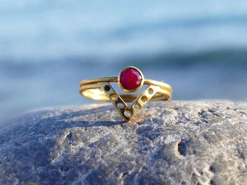 May include: A gold ring with a red gemstone set in a V-shaped band. The ring is on a gray rock.