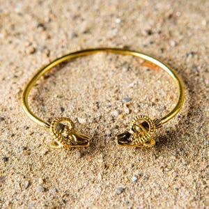 Könnte beinhalten: Ein goldfarbenes, offenes Armreif-Armband mit Widderkopfdetails an jedem Ende, das auf einer sandigen Oberfläche liegt. Das Armband hat ein kreisförmiges Design und ist wahrscheinlich aus Metall gefertigt. Die Widderköpfe sind aufwendig gestaltet.