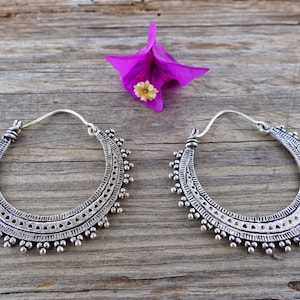 Puede incluir: Un par de pendientes de aro de plata con intrincados diseños geométricos. Los pendientes están hechos de metal y tienen una forma circular.