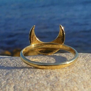 May include: A gold crescent moon ring with a smooth, polished finish. The ring is shown on a light-colored rock with a blue background.