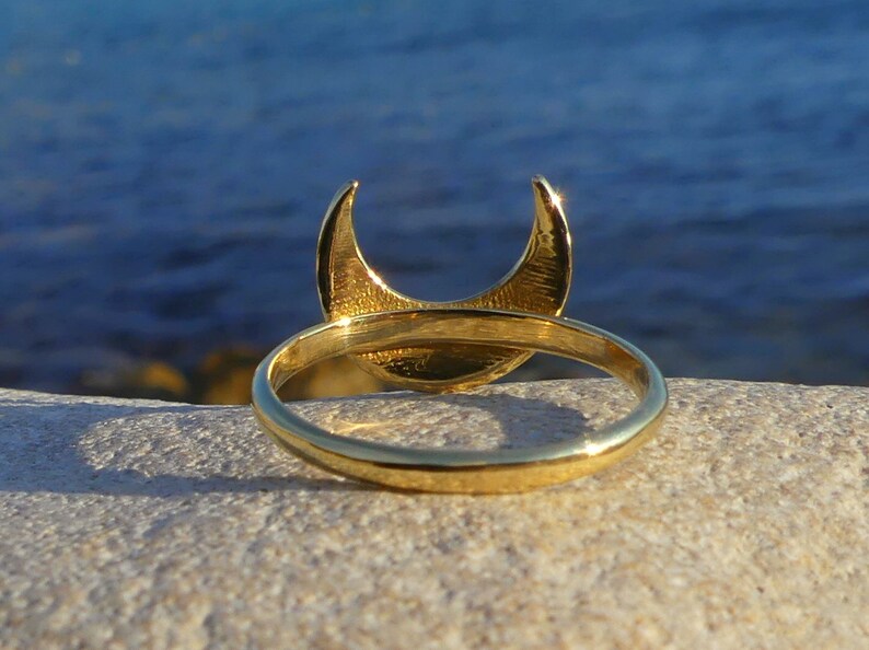 May include: A gold crescent moon ring with a smooth, polished finish. The ring is shown on a light-colored rock with a blue background.