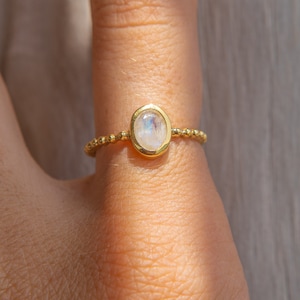 May include: A gold ring with a white oval moonstone set in a bezel setting. The ring is made of small gold beads.