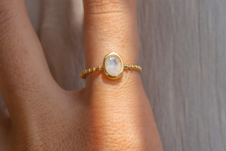 May include: A gold ring with a white oval moonstone set in a bezel setting. The ring is made of small gold beads.
