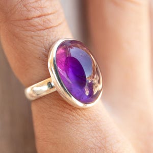 K&ouml;nnte beinhalten: Ein Silberring mit einem gro&szlig;en, ovalen Amethyst-Edelstein. Der tiefviolette Stein ist in einem polierten Silberband gefasst und schafft ein elegantes Schmuckst&uuml;ck. Der Ring ist ein Statement-St&uuml;ck, perfekt, um einen Farbtupfer zu setzen.