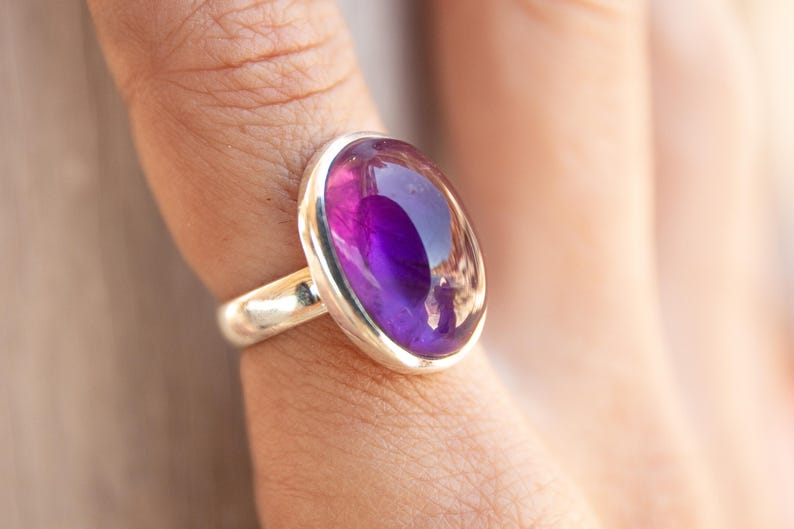 K&ouml;nnte beinhalten: Ein Silberring mit einem gro&szlig;en, ovalen Amethyst-Edelstein. Der tiefviolette Stein ist in einem polierten Silberband gefasst und schafft ein elegantes Schmuckst&uuml;ck. Der Ring ist ein Statement-St&uuml;ck, perfekt, um einen Farbtupfer zu setzen.