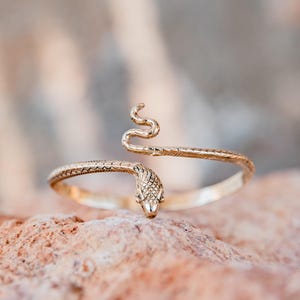 Puede incluir: Un brazalete de serpiente dorado. El brazalete presenta un diseño detallado de cabeza y cola de serpiente, con el cuerpo de la serpiente envolviendo el brazo. La cabeza de la serpiente está en la parte inferior y la cola se curva hacia arriba. El brazalete está sobre un fondo borroso.
