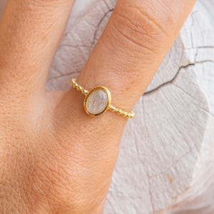 May include: A gold ring with a white oval gemstone. The ring is on a finger.