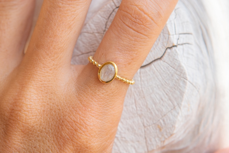 May include: A gold ring with a white oval gemstone. The ring is on a finger.