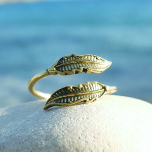 Puede incluir: Un anillo de tono dorado con dos diseños en forma de hoja. El anillo es abierto y ajustable.