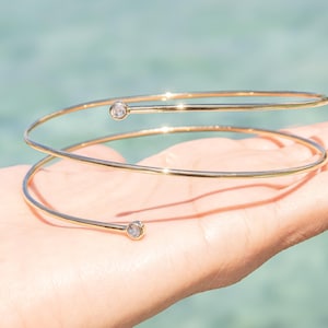 May include: A gold-toned spiral bracelet featuring a thin, polished metal band that wraps around in a double-loop design. Each end of the wire is finished with a small, round, clear stone setting, creating a simple and continuous circular silhouette.