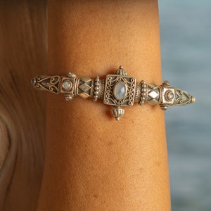 K&ouml;nnte beinhalten: Silberarmband mit aufwendigem Design. Das Armband hat ein zentrales Quadrat mit einem Mondstein und wird von dekorativen Elementen flankiert. Das Armband wird an einem Arm getragen, mit einer Holzoberfl&auml;che im Hintergrund.