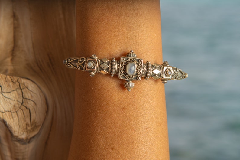 K&ouml;nnte beinhalten: Silberarmband mit aufwendigem Design. Das Armband hat ein zentrales Quadrat mit einem Mondstein und wird von dekorativen Elementen flankiert. Das Armband wird an einem Arm getragen, mit einer Holzoberfl&auml;che im Hintergrund.