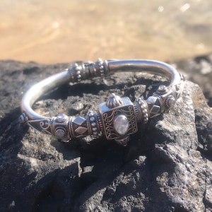 K&ouml;nnte beinhalten: Ein silberfarbenes Armband mit aufwendigen Details, das ein zentrales quadratisches Element mit einem klaren Stein aufweist. Das Armband liegt auf einem dunklen, strukturierten Felsen mit verschwommenem Wasserhintergrund, was sein kompliziertes Design und seine Handwerkskunst zeigt.